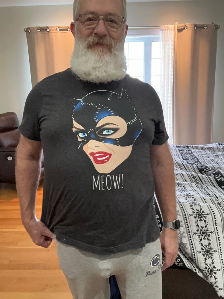 Gray-bearded man wearing a Catwoman T-shirt and gray sweat pants with visible penis outline showing in the sweats.