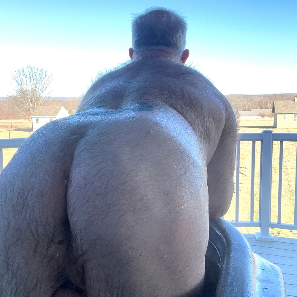 Naked hairy dude leaning over the side of his hot tub, butt exposed, looking into the back yard.