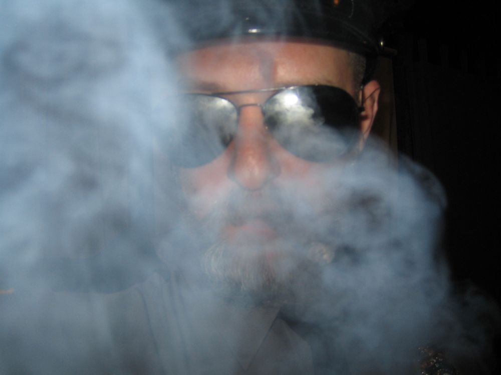 Smoke obscures most of the face of a man wearing mirrored sunglasses.