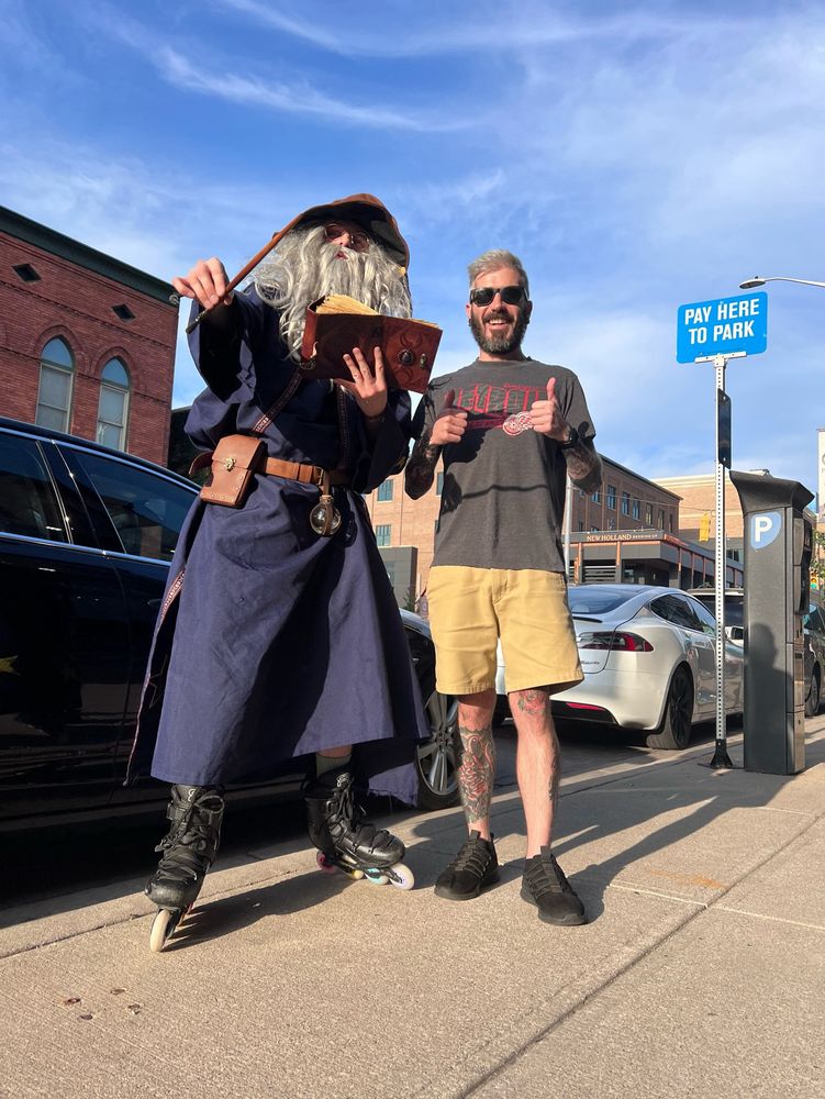 A photo of a man who dresses fully as a wizard on rollerblades next to the poster giving a thumbs up on a city block on a sunny day 