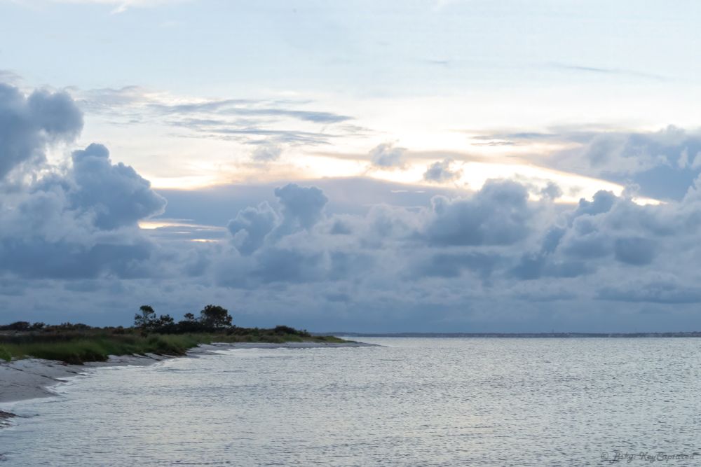 A wide landscape shot of a sunset behind some dense clouds over the sound in the horizon. Closer to the viewer, the sparse clouds above can be seen under lit by the setting sun, soon to be overtaken by the approaching rain.