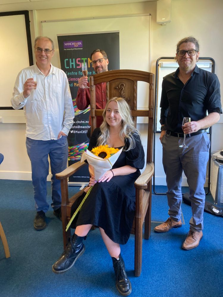 Photograph of a young woman holding sunflowers sitting in a throne-like chair, with three men standing behind her holding champagne glasses.