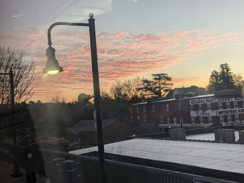 Photo through a slightly smudged window of a decorative streetlight, a train platform, and in the background a brick warehouse with the words Gunston Hall, Janney Hall, Coffee Importers visible. The sunrise is turning some wispy clouds orange.