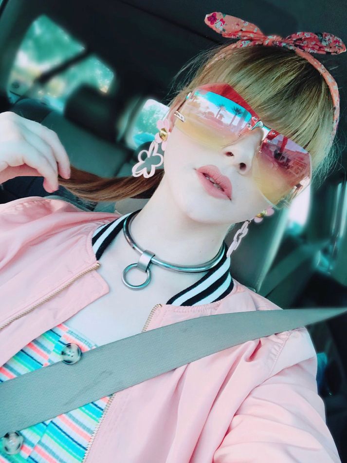 A blonde, transgender woman sitting in the passenger seat of a car. She’s is wearing a striped top, a pink bomber jacket and outrageous peach & yellow sunglasses. The streets of Las Vegas are reflected in her glasses. 