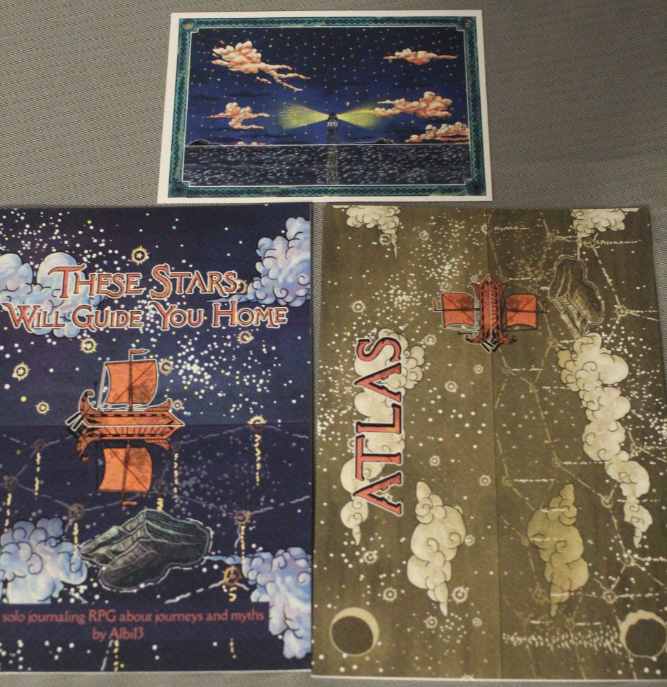 a picture of the These Stars Will Guide You Home zine (night sky background, with constellations, and a lonely orange ship sailing at the front, near a small island), along with the Atlas zine (night sky background, but in lighter tones, filled with constellations and the same boat and island). also, an island print depicting a star-filled night sky, some clouds and a lighthouse.