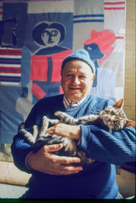 A color photo of the artist Romare Bearden, a large work behind him, cradling his cat, Gippo, who looks back at us