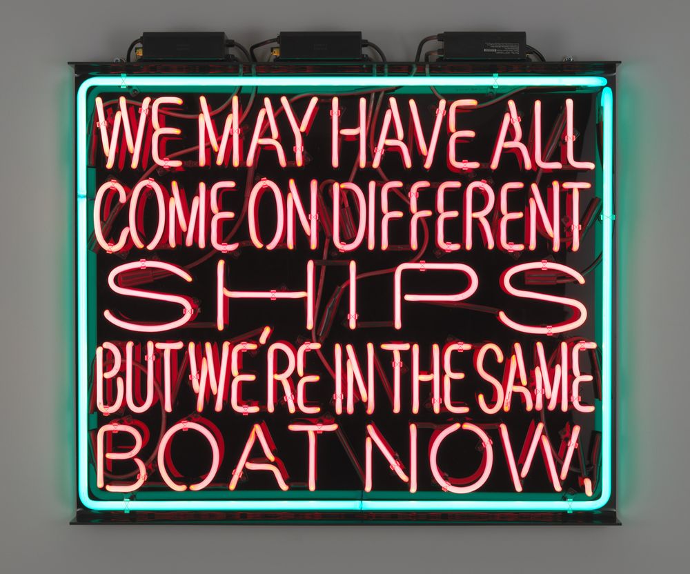 Neon sign outlined in green and reading in pinkish-red letters, "We may have all come on different ships but we're in the same boat now."