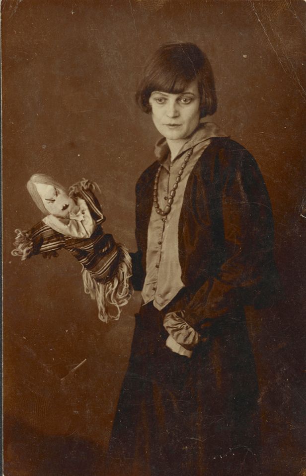 Sepia tinted photo of a woman, almost full-length, in a bob haircut (indicative of first decades of the 20th century) with a puppet - apparently of an angry or stern woman - on her right hand