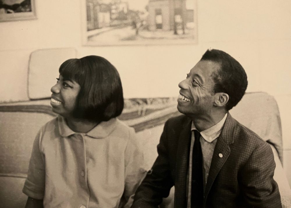 Photo of Nina Simone and James Baldwin, seated on a sofa or couch, looking up and to the left of the photo, both smiling broadly