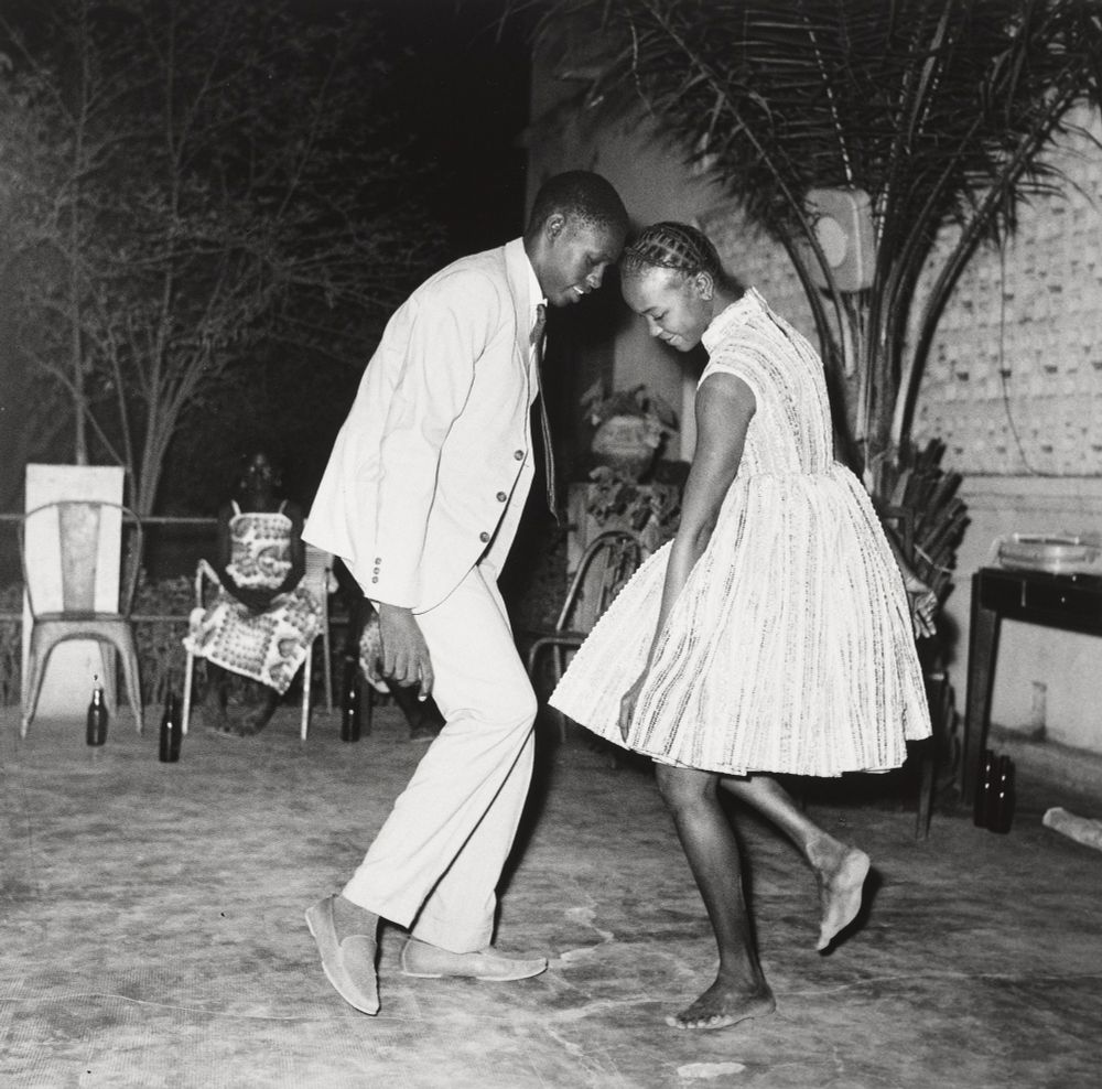 Two people, a man and a woman, dance on an open air dance floor. The two are close together, their heads almost touching. Both look down, apparently at their feet. This black and white photo conveys a palpable sense of joy