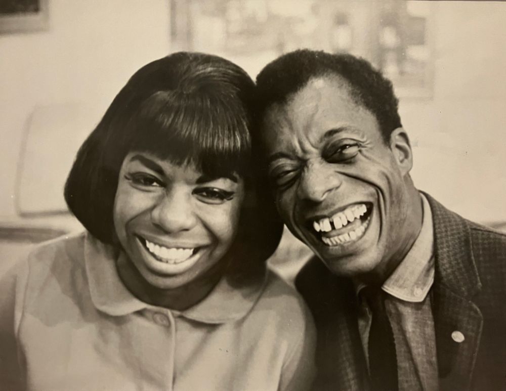 Photo of Nina Simone & James Baldwin. They have their heads together, looking out at us and smiling and laughing broadly, as if having the times of their lives. An image of pure JOY
