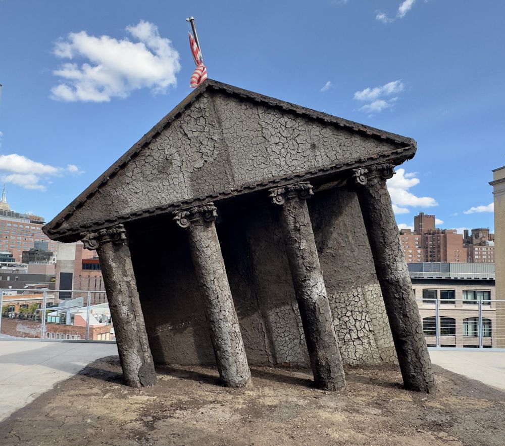 Sculpture of a dark, cracked classical architectural facade listing as if collapsing, with American flag above