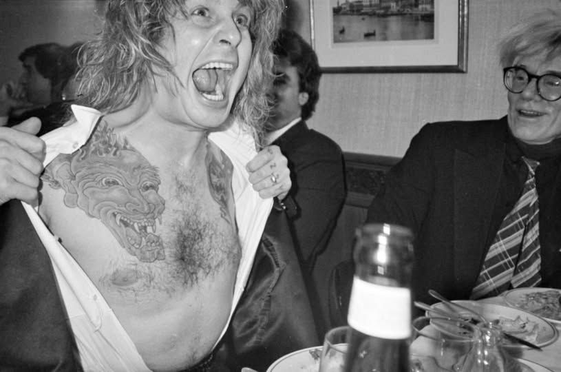 B&W photo of Ozzy Osbourne seemingly screaming and pulling open his shirt, with Andy Warhol looking on apparently bemused at right