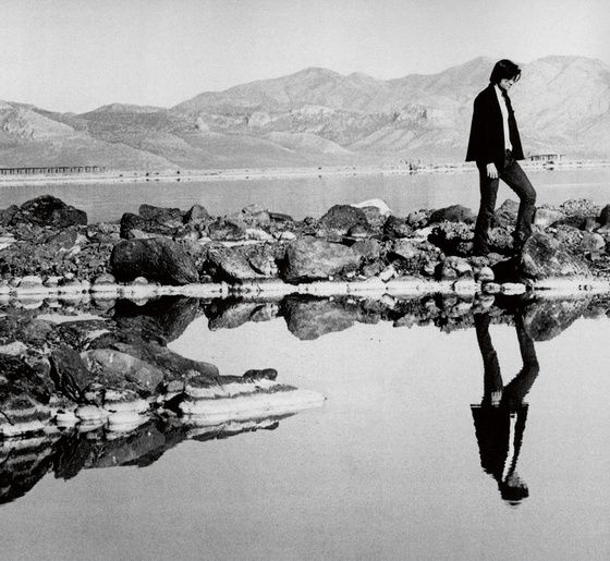 B&W photo of man walking on rocks, his reflection in water below him