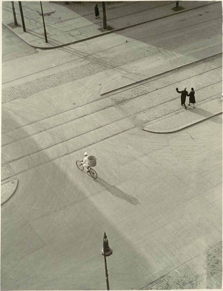 Photograph shot from above, aka a bird's eye view, of an urban street with tram tracks, and three figures: a delivery man (perhaps for bread?0 on a bicycle, and a male and female couple walking arm in arm. The long shadows suggest it is early in the morning