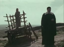 A cloaked figure stands in front of a gallows being built 