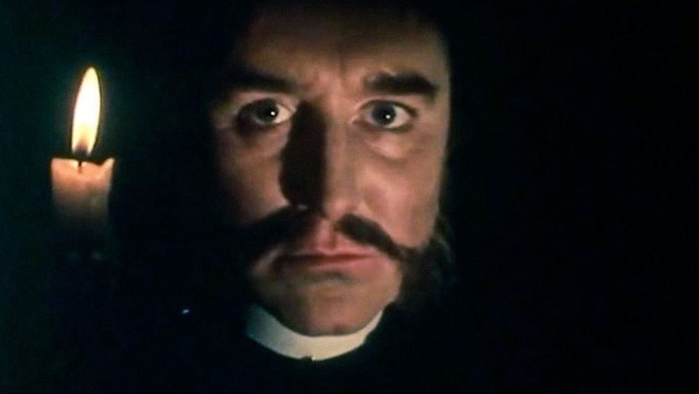 Robert Hardy as the Archdeacon facing the camera holding a candle up near his face as he stands in pitch darkness.