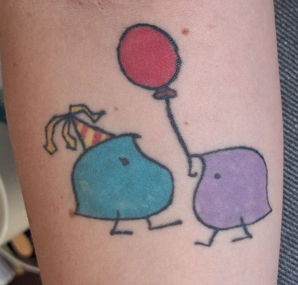 A tattoo of two wugs having a party. The one on the left is blue and is wearing a yellow and red striped party hat. The one on the right is purple and is holding a red balloon.