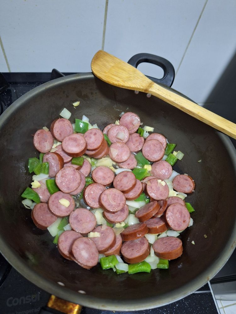 Cebola, alho e pimentão verde cortados em uma panela grande junto com linguiça calabresa fatiada sendo fritas em óleo quente.
