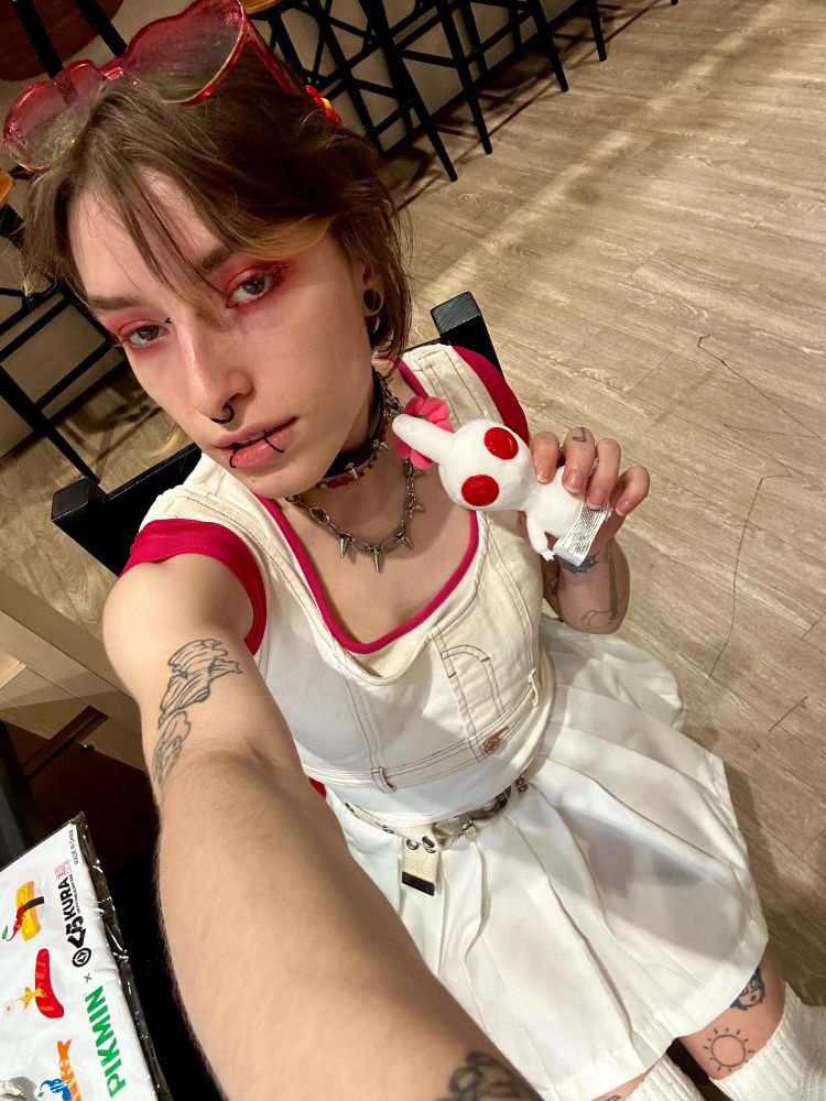 i am taking a selfie sitting in a chair holding a white pikmin. i am wearing a white outfit with red accents with heart sunglasses and have on red eye makeup