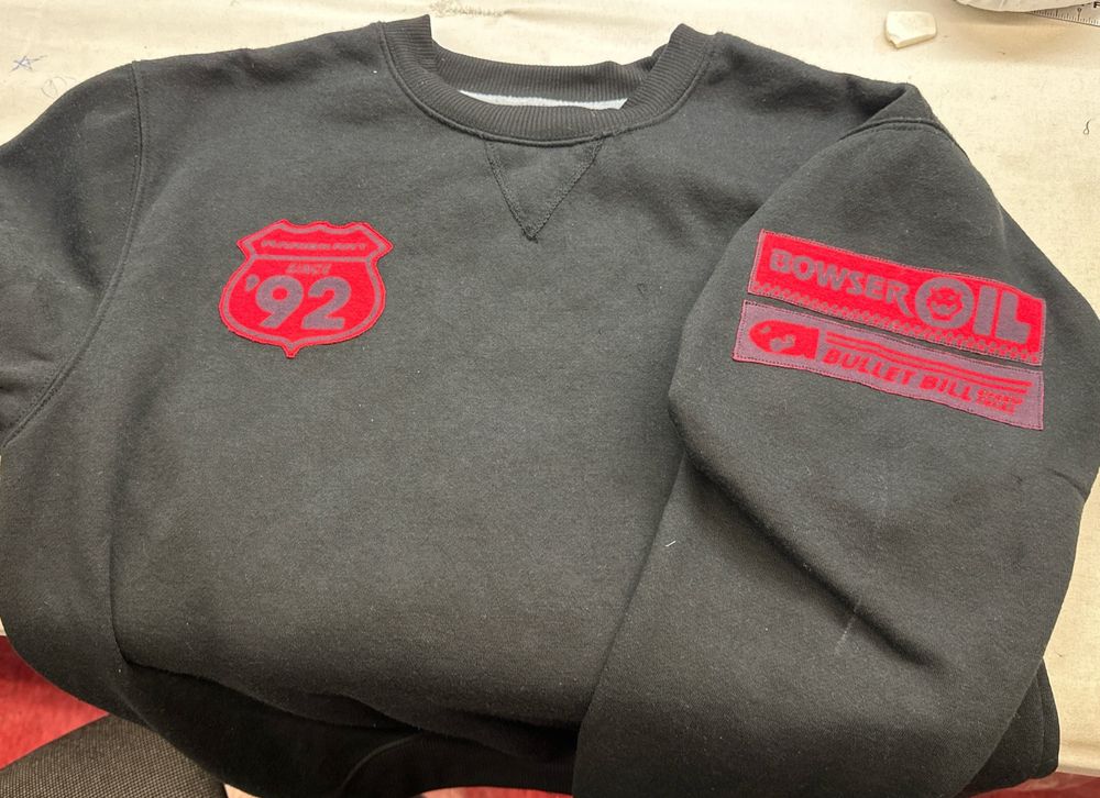 black sweatshirt laid out on a table with bright red mario kart graphics. the graphic over the left breast reads "mariokart since '92" in the shape of a road sign. the two recangular graphics centered on the top of the outer right sleeve read "bowser oil" and "bullet bill speed trial"