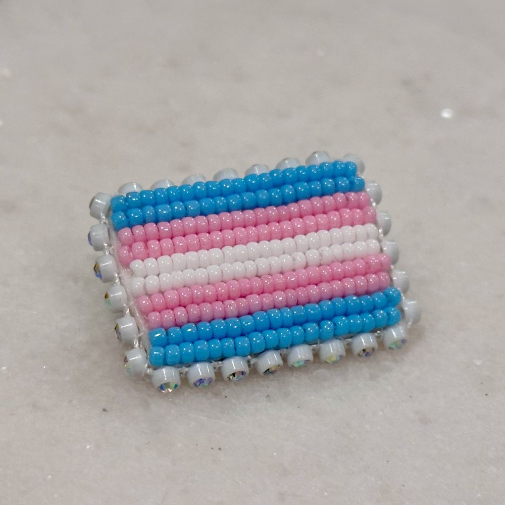 A beaded trans pride flag pin edged in rhinestones on a white background