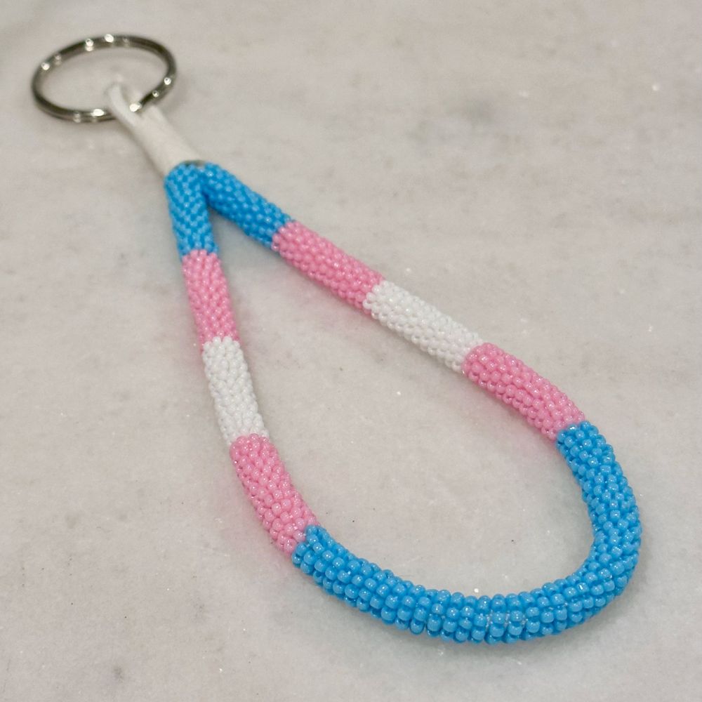 A bead wrapped keychain in the trans pride colors on a white marble background