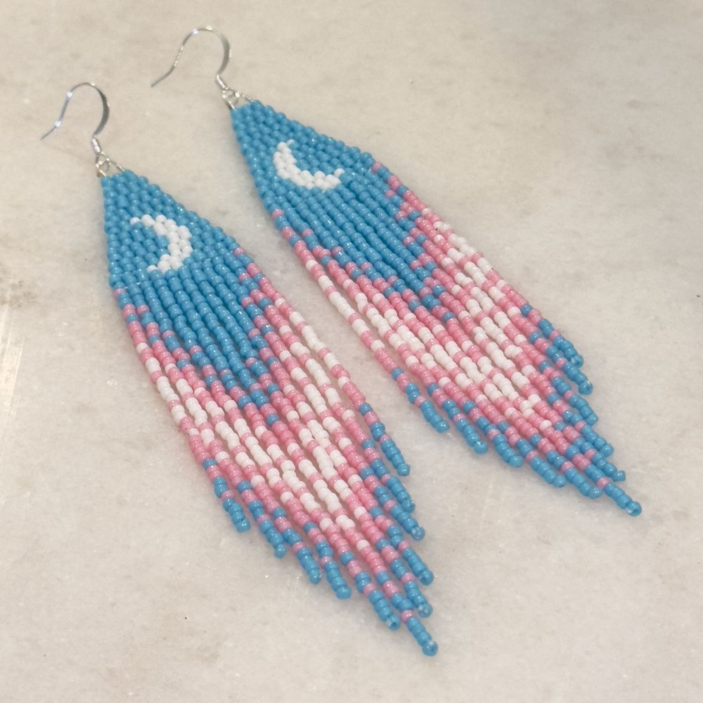 A pair of beaded fringe earrings in the trans pride colors with a white crescent moon shape on the brick stitch top