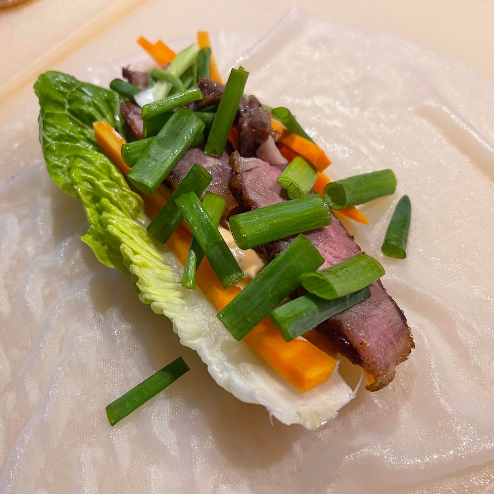 Na zdjęciu znajduje się arkusz papieru ryżowego, na którym leży liść sałaty rzymskiej, paski marchewki, kawałki różowego steku oraz posiekany szczypior.