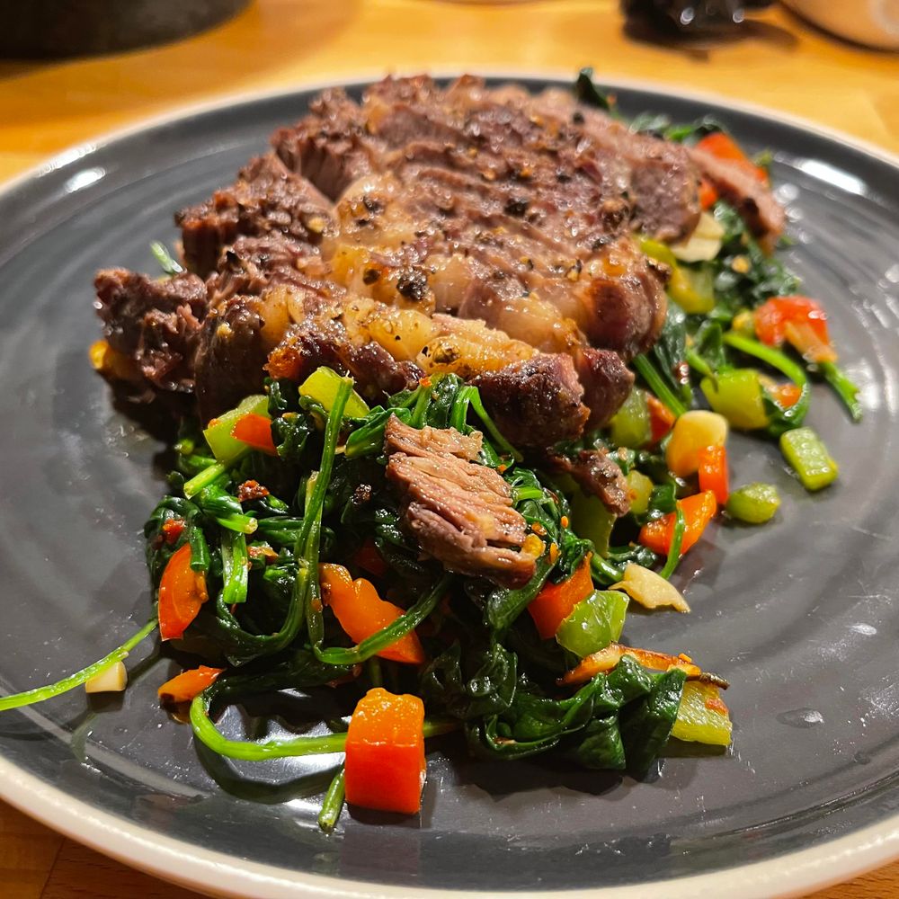 Na ciemnym talerzu leży pokrojony stek wołowy ułożony na blanszowanym szpinaku z kawałkami czerwonej i zielonej papryki. Danie stoi na drewnianym blacie kuchennym.