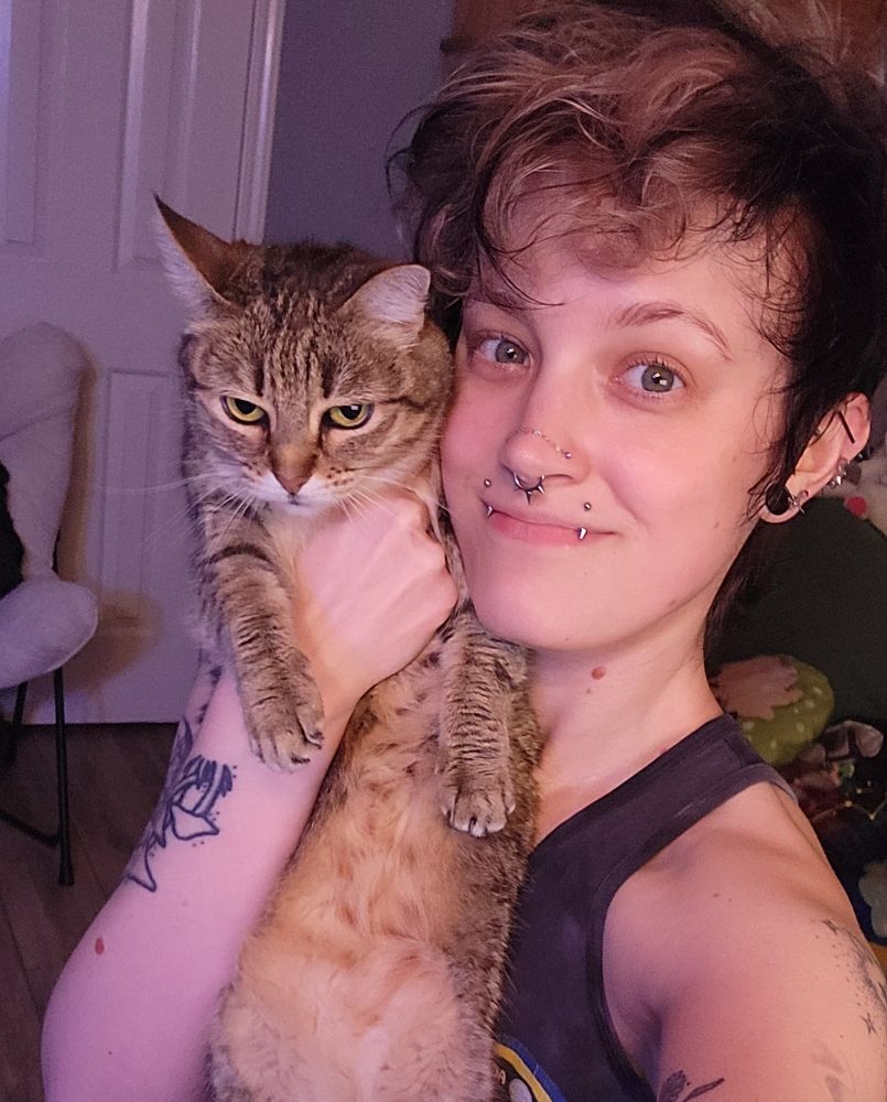 Person with short black and white hair holding up a annoyed looking tabby cat