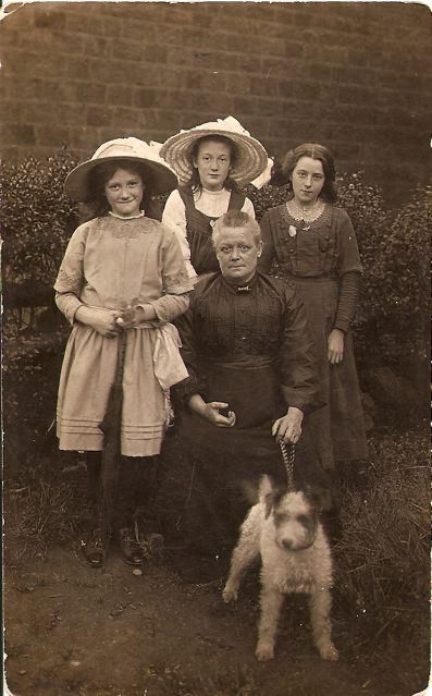 Mother seated in black dress with her three daughters standing behind and family dog in front 