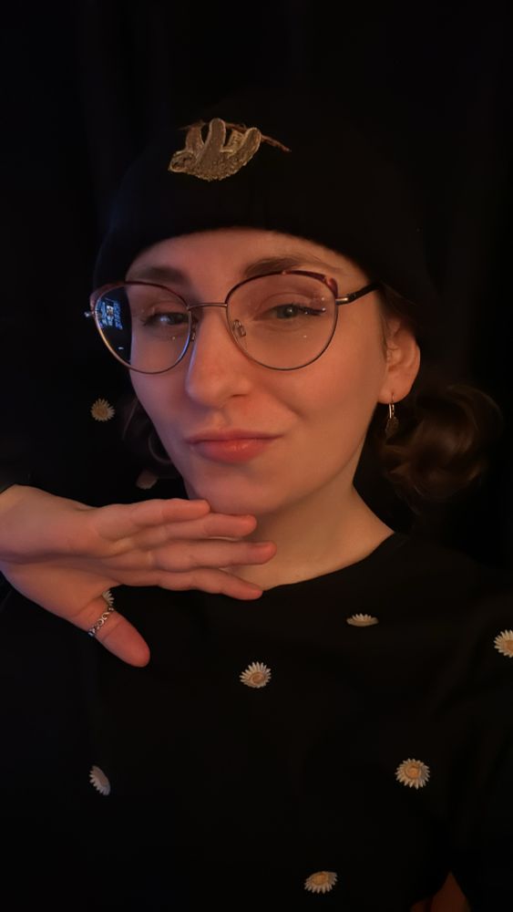 most recent selfie of Tasha, she is wearing glasses, a black beanie, hair in small but low space buns, little drop earrings with metal feathers and has her hand raised up, pointed and flat, under her chin.