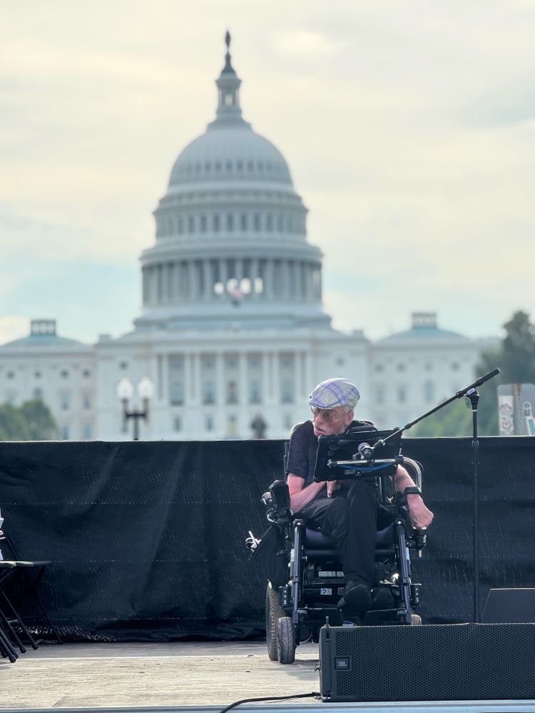 [Image: Bob Williams wears a plaid hat and glasses. He sits in a powerchair with a microphone near his speech-generating device. Behind him is the US Capitol building.]