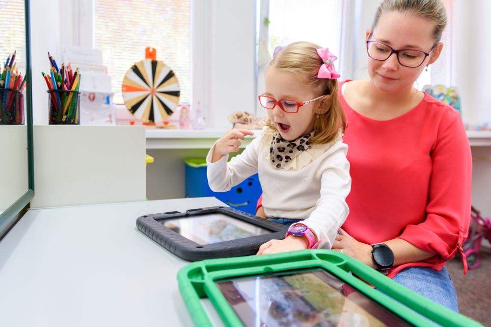 [Image: In a classroom, a woman and a young girl sit at a table with two tablets on it. They are focused on the tablet closest to them, which the girl is using.]