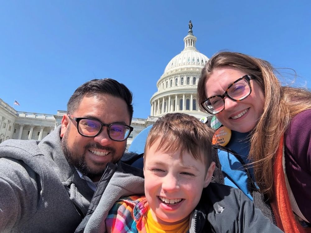 [Image: A selfie of Toby Latham, his wife Lindsay, and their young son who is an AAC user. With the U.S. Capitol and clear blue skies in the background, the three of them smile widely.]
