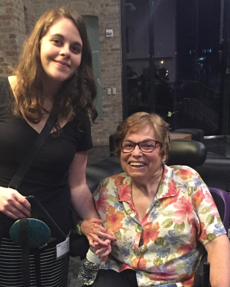 CommunicationFIRST Board Chair Jordyn Zimmerman, standing in black, holds hands with a smiling Judy Heumann, who sits in a power wheelchair wearing a floral shirt.