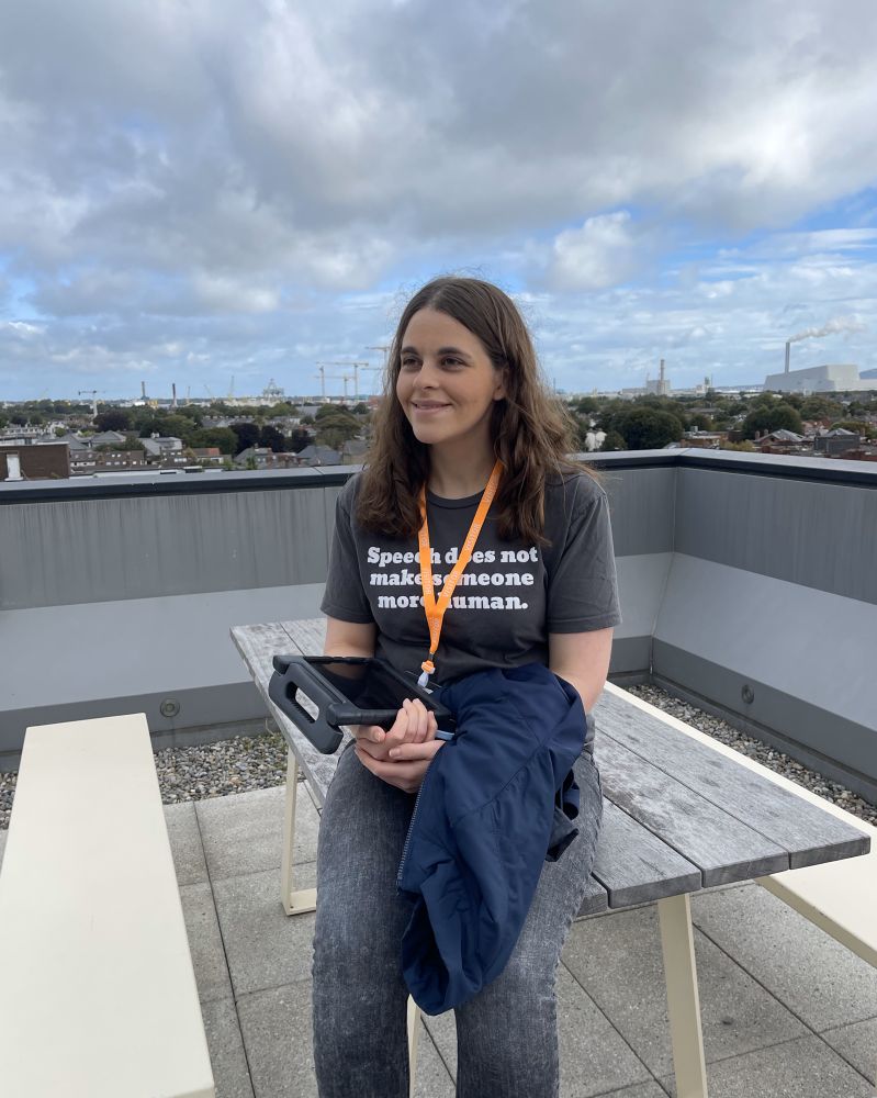 [Image: Jordyn Zimmerman sits smiling on a rooftop bench, wearing a shirt that says “Speech does not make someone more human.” She holds an AAC device and a jacket. Behind her, partly cloudy skies and a tree-lined cityscape stretch into the distance.]
