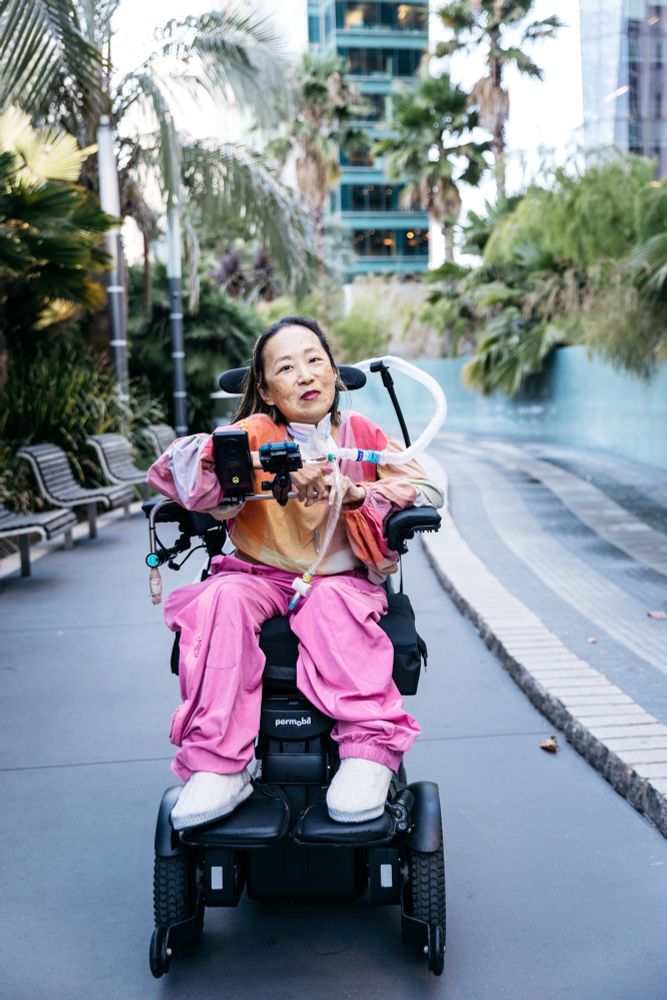 [Image: Alice Wong in her wheelchair with a tracheostomy at her neck connected to a ventilator. A smiling Alice wears a pink plaid shirt and pink pants, and her AAC device is mounted in front of her.]
