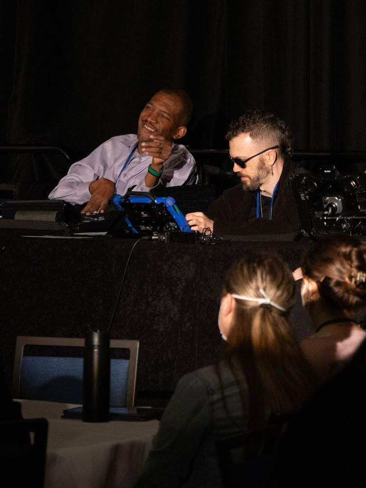 [Image: AAC users Lateef McLeod (left) and Lance McLemore (right) present on stage at the Future of AAC Research Summit in May 2024. Lateef and Lance type on communication devices. Across from them, the backs of two audience members’ heads are visible. They appear to be listening attentively]. 
