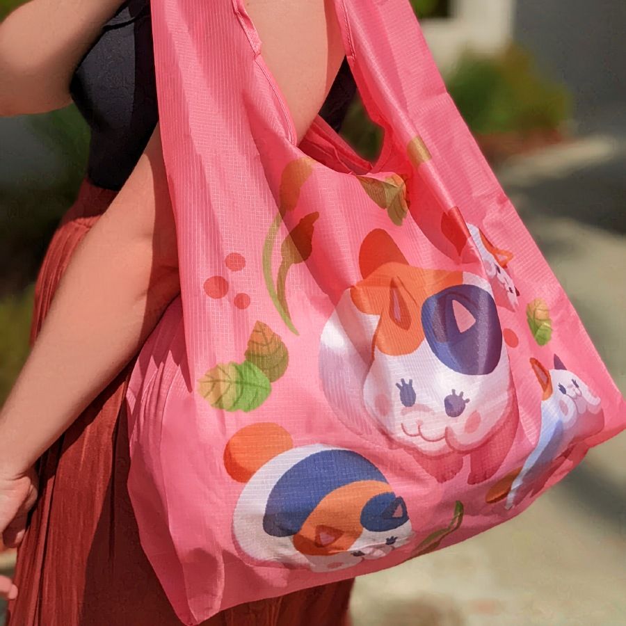 Product photo of myself carrying a pink, eco-friendly, Final Fantasy XIV-inspired Fat Cat bag over my shoulder. All bags are made of ripstop nylon and the carrying portion is roughly 16" x 16" with gusseted sides allowing for expansion up to 21" wide.