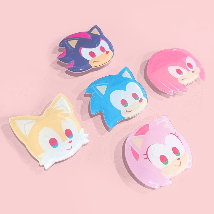 Product photo of 5 Sonic phone grip designs—Sonic, Tails, Knuckles, Amy, and Shadow—arranged in a neat cross against a pink backdrop. Each design measures approximately 6 x 5 centimeters, and they are made of acrylic.