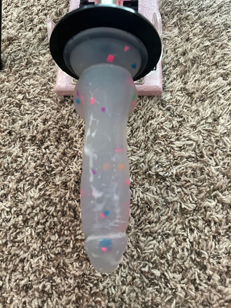 A clear dildo with rainbow confetti is attached to a pink fuck machine. There is a creamy liquid all over the dildo 
