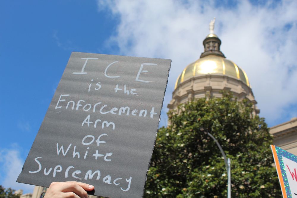 A protester holds a sign reading “ICE is the enforcement arm of white supremacy” with Georgia’s Gold Dome Capitol building in the background. 