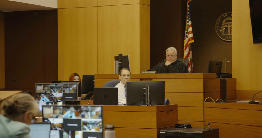 A courtroom. Judge Kevin M. Farmer sits in the back of the frame, presiding over the trial. A court reporter and clerk are visible in front and to the side of the judge. 

A prosecutor and screens showing the courtroom cameras sit in the foreground.
