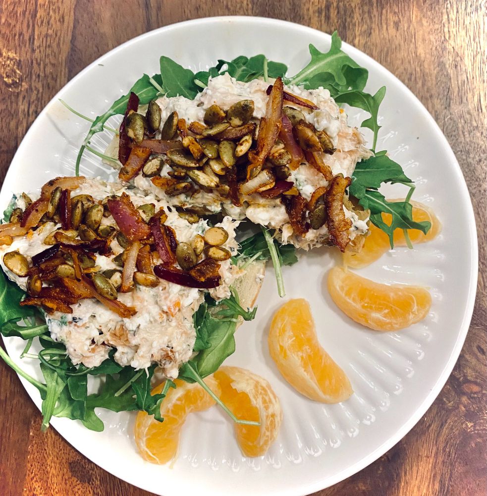 A toasted parmesan bagel topped with creamy herbed cottage cheese spread, fresh arugula, flaked salmon, and smoky spiced pepitas sautéed with caramelized red onions. Pepitas glisten with smoked paprika and ground mustard seasoning, while bright orange tangerine slices sit alongside, adding a pop of color and citrusy freshness.

Recipe:
1.	Herbed Cottage Cheese Spread: Blend 1/2 cup cottage cheese, 1 tbsp heavy cream, and 1 tsp lemon juice. Fold in 1 tbsp chopped chives, 1 tsp dill, 2-3 chopped green olives, and 1/2 tbsp minced red onion. Season with salt & black pepper.
2.	Spiced Pepitas: In a skillet, toast 2 tbsp pepitas with a drizzle of oil (or water), 1/4 tsp smoked paprika, 1/4 tsp ground mustard, and a pinch of salt. Add 1/2 tbsp minced red onion; sauté until the pepitas are golden and the onions caramelize.
3.	Assemble:
•	Toast a parmesan bagel.
•	Add a handful of arugula.
•	Spread the herbed cottage cheese generously.
•	Top with flaked salmon.
•	Finish with the spiced pepitas and onions for flavor and crunch.