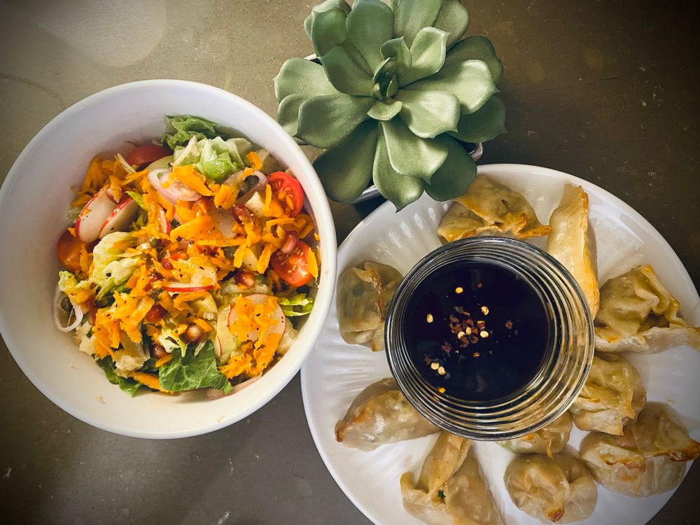 oven-baked shrimp gyoza and a vibrant, refreshing salad. The gyoza are arranged in a circular pattern on a white plate, with a small glass dish of dipping sauce in the center. The sauce is dark, garnished with a sprinkle of red pepper flakes for a pop of color. The gyoza have a lightly golden exterior, with some pleated and others folded into crescent shapes, showcasing their handcrafted nature.

Beside the gyoza plate is a fresh, colorful salad served in a white bowl. The salad includes chopped romaine lettuce and Napa cabbage, shredded carrots and freshly grated ginger, diced grapefruit, pomegranate arils, halved cherry tomatoes, chopped artichoke hearts, and capers, all tossed in G Hughes Sugar-Free Asian Miso Dressing. The combination of textures and flavors—sweet, tart, briny, and fresh—makes for a well-balanced and satisfying dish.

📌 Oven-Baked Shrimp Gyoza Recipe

Filling:
	•	½ lb shrimp, minced
	•	½ to ¾ cup Napa cabbage, finely chopped & squeezed dry
	•	2 green onions, finely chopped
	•	¼ cup carrots, shredded
	•	½ to ¾ tsp freshly grated ginger
	•	¼ tsp ground garlic
	•	2 tbsp liquid aminos
	•	1 tsp rice vinegar
	•	Pinch of white pepper (optional)

For Baking:
	•	Nonfat avocado cooking spray
	•	1 small ramekin of boiling water

Dipping Sauce:
	•	2 tbsp liquid aminos
	•	1 tsp rice vinegar
	•	1 tsp toasted sesame oil
	•	Optional: chili flakes or hot honey

Instructions:
	1.	Mix all filling ingredients in a bowl until well combined.
	2.	Place 1 tsp of filling in a gyoza wrapper, wet edges, fold, and pleat to seal.
	3.	Preheat oven to 400°F (204°C). Line a small roasting pan with aluminum foil, spray with nonfat avocado spray, and arrange gyoza in a single layer.
	4.	Place a ramekin of boiling water on the pan to create steam. Lightly mist the gyoza with avocado spray.
	5.	Bake for 10 minutes, mist again if needed, and bake for another 5-10 minutes until lightly golden. Broil for 1-2 minutes for extra crispness.
	6.	Serve with dipping sauce.
