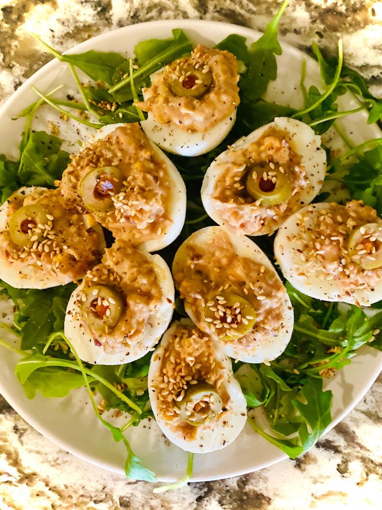 A plate of cranberry mustard deviled eggs arranged on a bed of fresh arugula. The eggs are sliced in half lengthwise, revealing a creamy and textured filling made by mashing hard-boiled yolks with cranberry mustard, a pinch of garlic powder, black pepper to taste, nutritional yeast, and diced green olives. Each deviled egg is topped with a sliced green olive, its center filled with pimento, and a light sprinkle of sesame seeds for extra texture. The eggs are garnished with freshly ground black pepper, and the arugula underneath adds a vibrant, green contrast to the dish.

This recipe uses six large hard-boiled eggs, peeled and halved. The yolks are scooped into a bowl and combined with two tablespoons of cranberry mustard, one teaspoon of nutritional yeast, two tablespoons of finely diced green olives, a pinch of garlic powder, and black pepper to taste. The mixture is mashed until smooth and creamy, then spooned back into the egg whites. Each egg is garnished with a slice of green olive and sprinkled with sesame seeds for crunch. The presentation features the eggs arranged in a circular pattern on a plate with fresh arugula, creating an elegant and colorful visual.

The cranberry mustard adds a tangy and slightly sweet note to the rich egg yolk, balanced by the briny flavor of the green olives. Nutritional yeast provides a subtle umami depth, while the sesame seeds add texture. The arugula acts as both a presentation element and a peppery, fresh complement to the dish.

This dish was created as a thoughtful response to a day when comfort and nutrition were needed, showcasing how food can be both medicine and art. The combination of flavors and textures elevates traditional deviled eggs into a unique and nourishing meal.