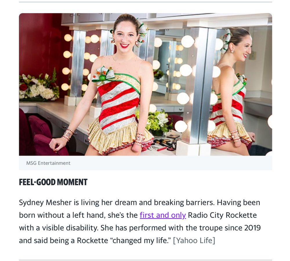 A young woman wearing a festive candy cane-themed costume with gold fringe sits in a dressing room with large, illuminated mirrors. She is smiling confidently. Born without a left hand, she is the first Radio City Rockette with a visible disability, breaking barriers in performance arts.