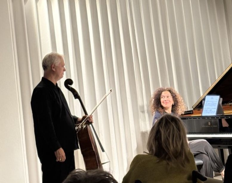 Man standing with a cello in hand looks at woman sitting at a grand piano with her head turned towards him.